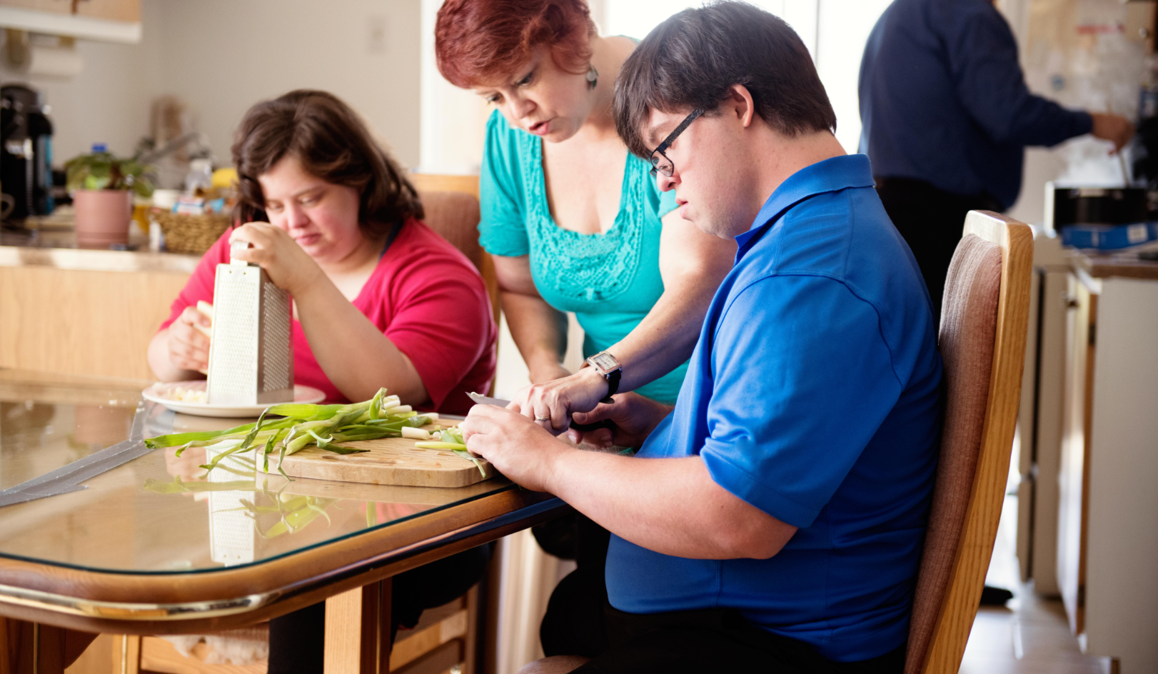 A middle aged woman helping two young adults seated at a kitchen table chop food.